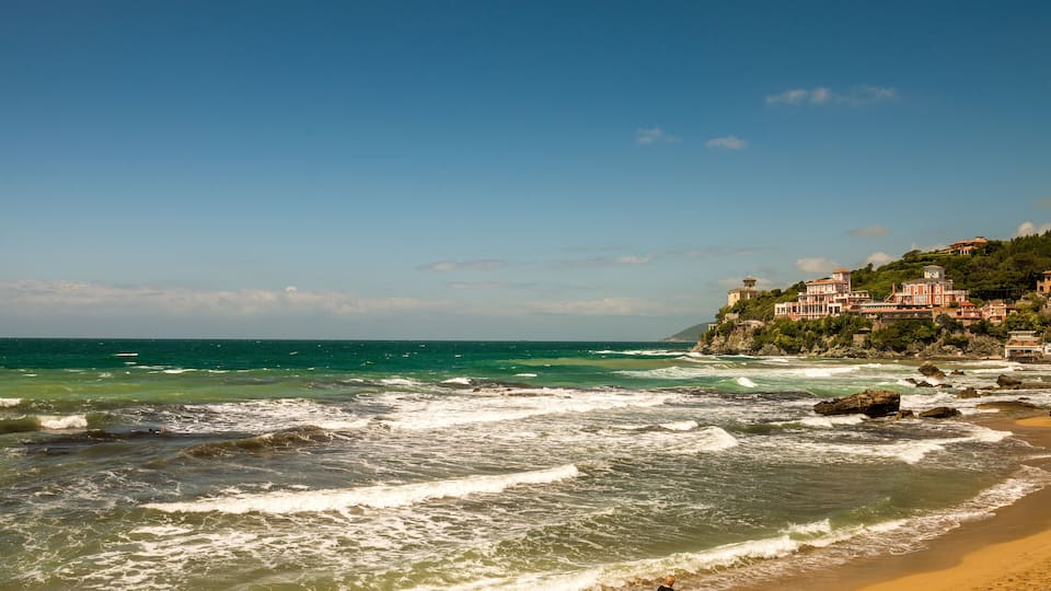 A beach with waves, a person walking, and buildings on a hill.