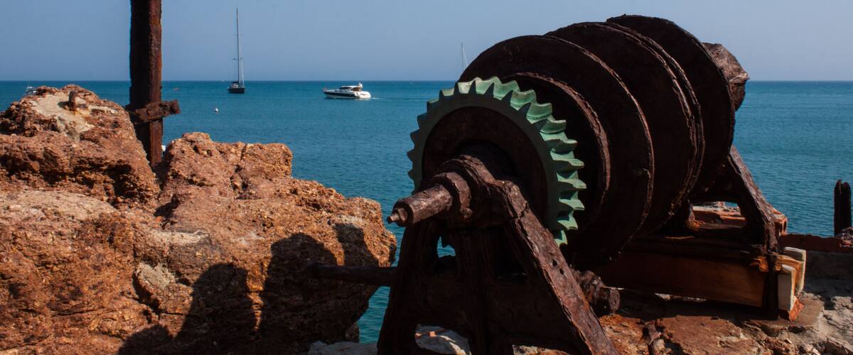 A rusty metal gear and wooden structure by the sea with boats in the background.