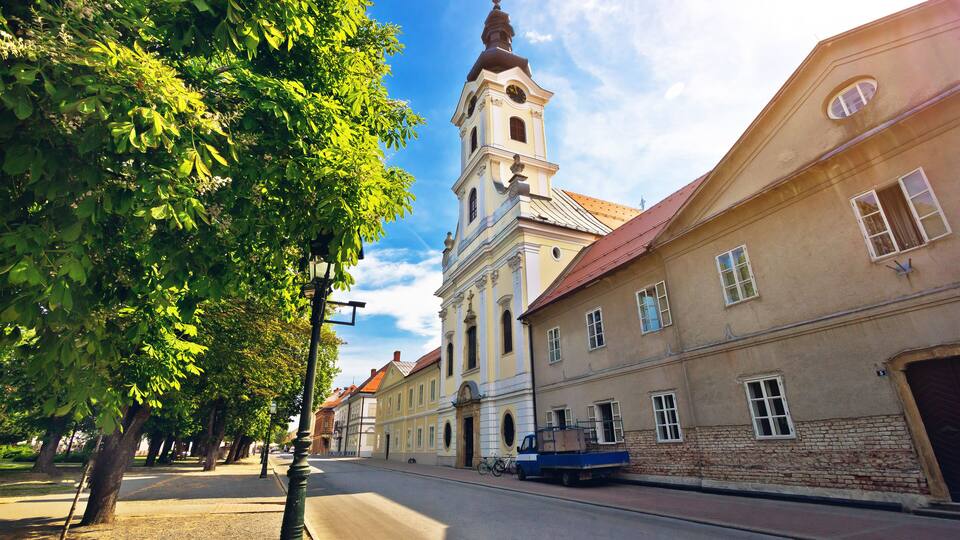 A street with a church tower, trees, and a blue truck.