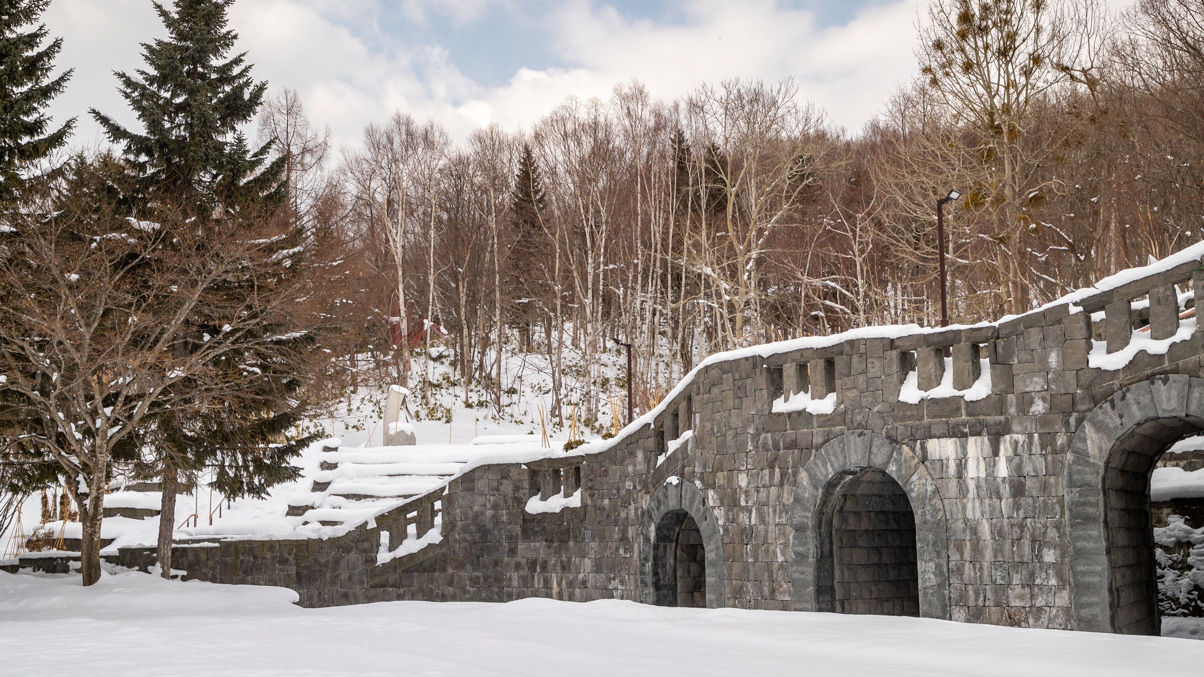 A stone bridge with snow-covered steps and arches, surrounded by trees and a forested hillside.