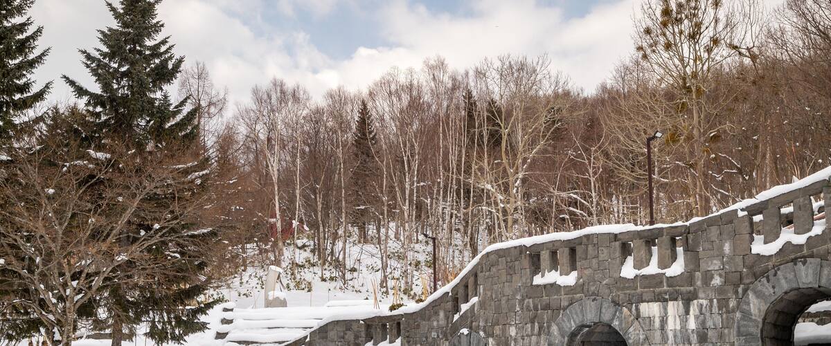 A stone bridge with snow-covered steps and arches, surrounded by trees and a forested hillside.