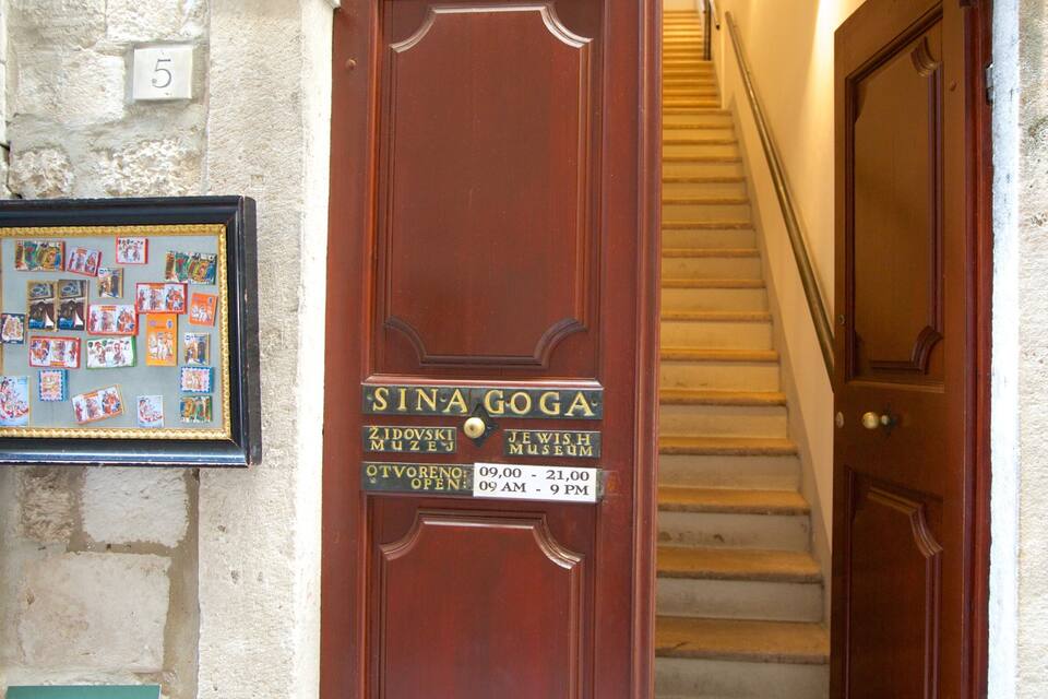 A wooden door with a sign for the Jewish Museum, a framed picture of postcards, and a staircase.