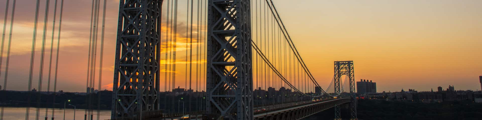 A large suspension bridge with a prominent arch and multiple cables, spanning a body of water with a city skyline in the background.