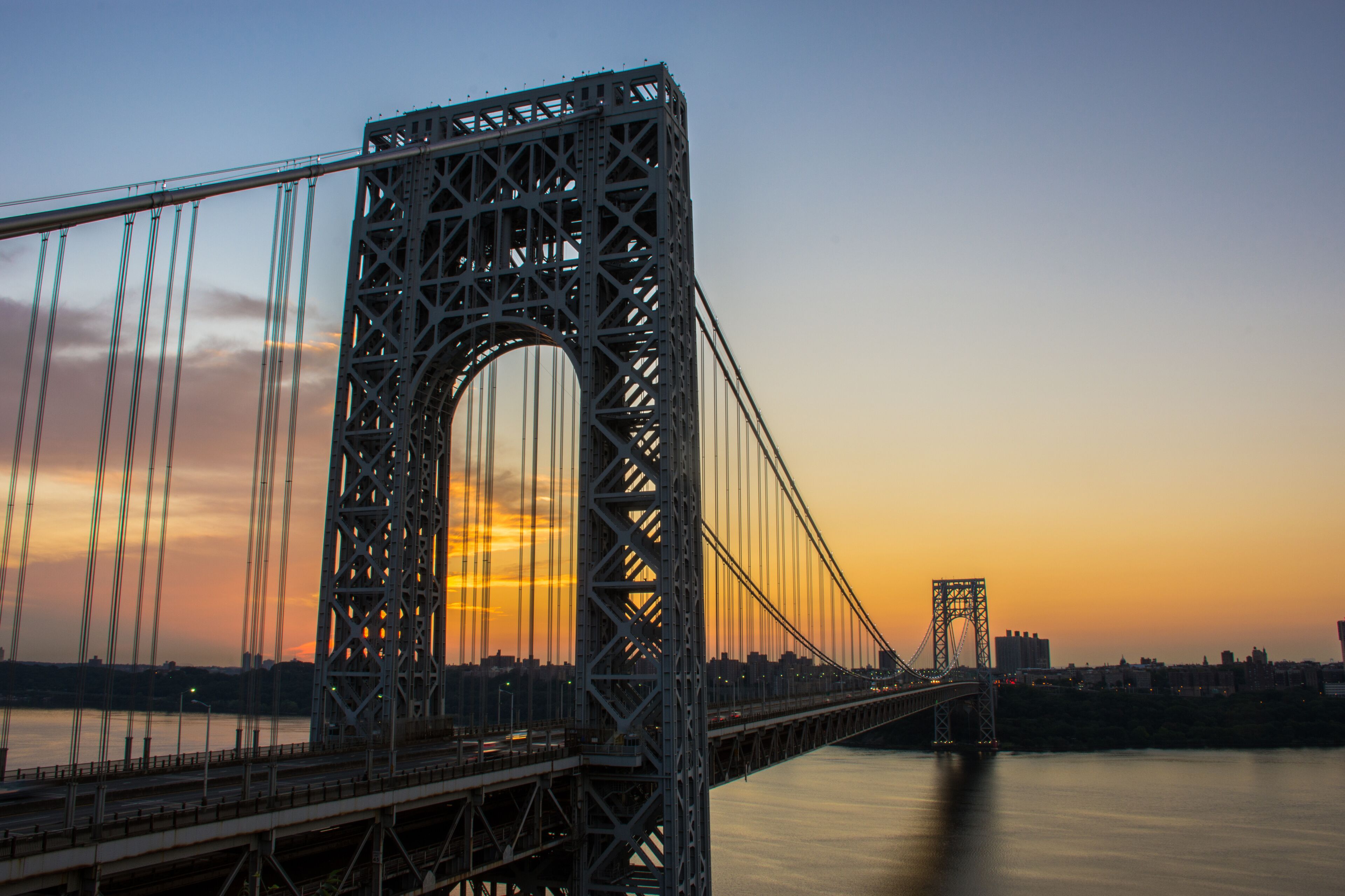 A large suspension bridge with a prominent arch and multiple cables, spanning a body of water with a city skyline in the background.