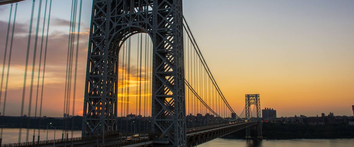 A large suspension bridge with a prominent arch and multiple cables, spanning a body of water with a city skyline in the background.