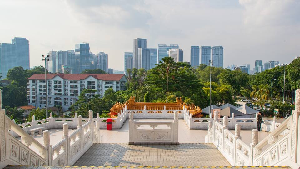 A traditional gate with ornate carvings, leading to a temple with golden statues. In the background, there are modern high-rise buildings and lush green trees.