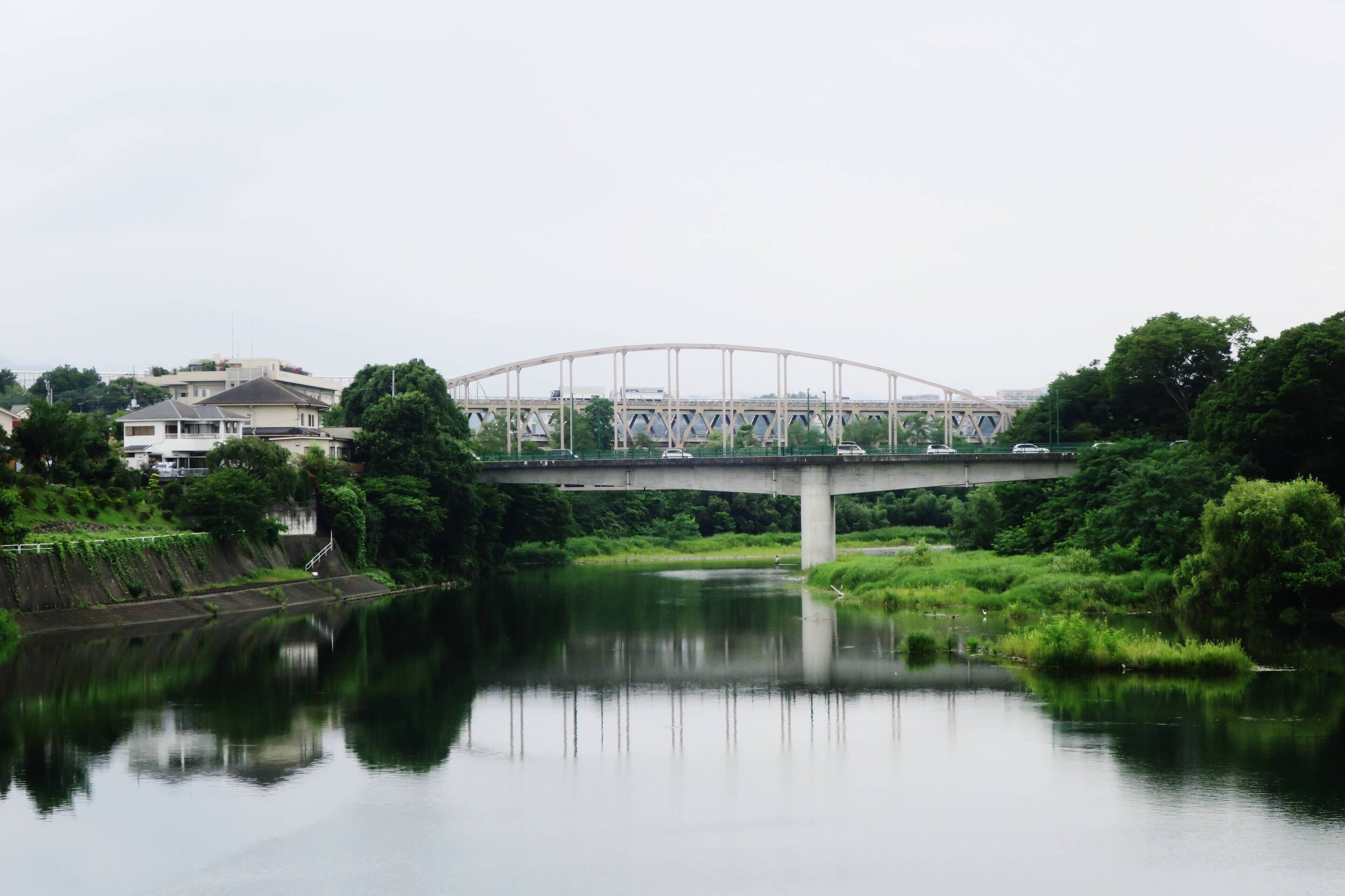 A bridge over a river with a reflection, surrounded by greenery and houses.