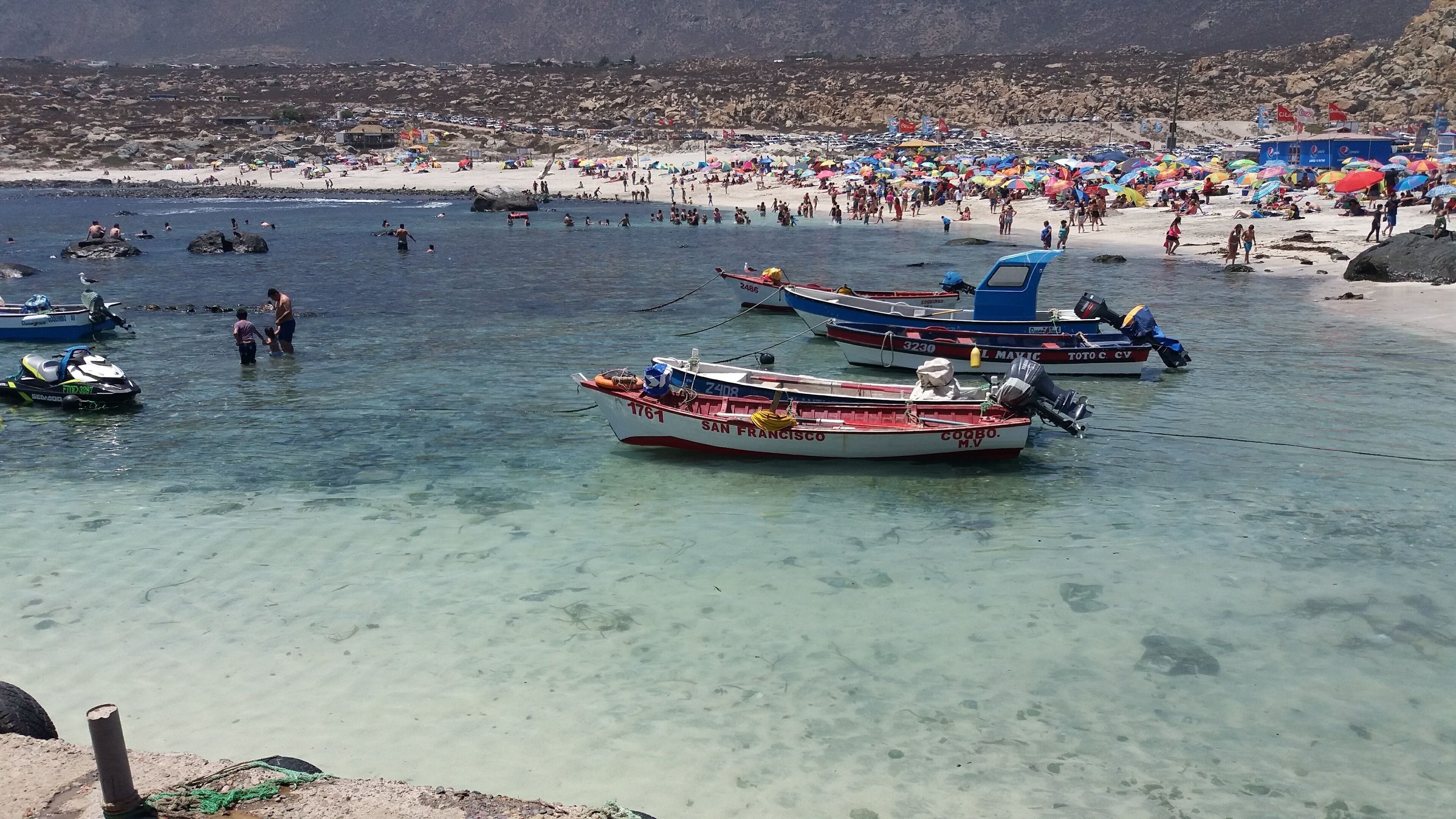 A beach with clear shallow waters, several boats anchored, and a crowded sandy shore.