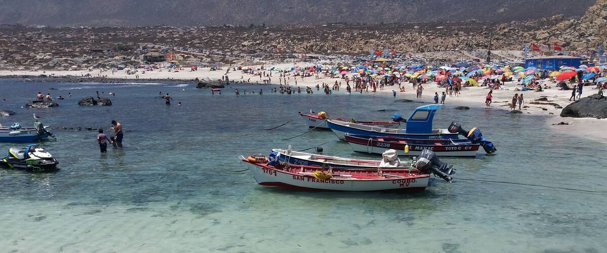 A beach with clear shallow waters, several boats anchored, and a crowded sandy shore.