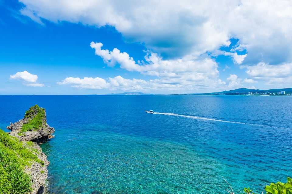 A coastal landscape with clear blue water, rocky outcrops, and lush greenery.