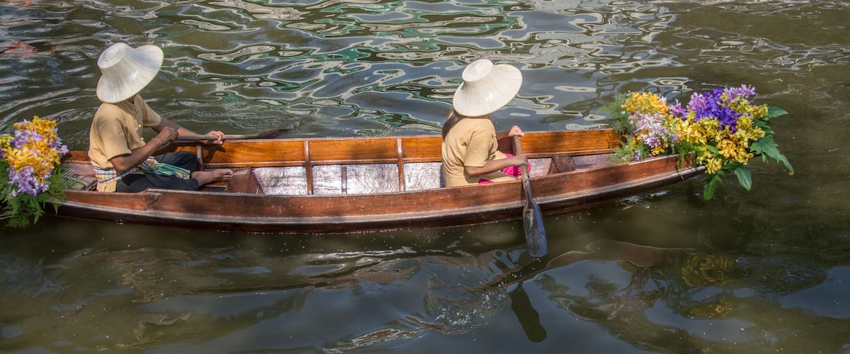 Two people in a wooden boat on a calm body of water, wearing conical hats and holding paddles.