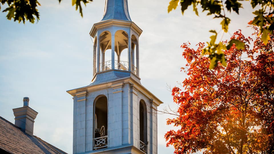 A church steeple with a bell tower and a red-leafed tree in the foreground.