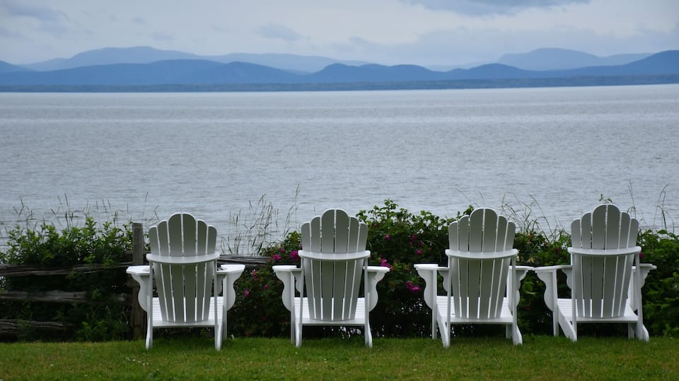 Three white Adirondack chairs facing a large body of water with mountains in the background.