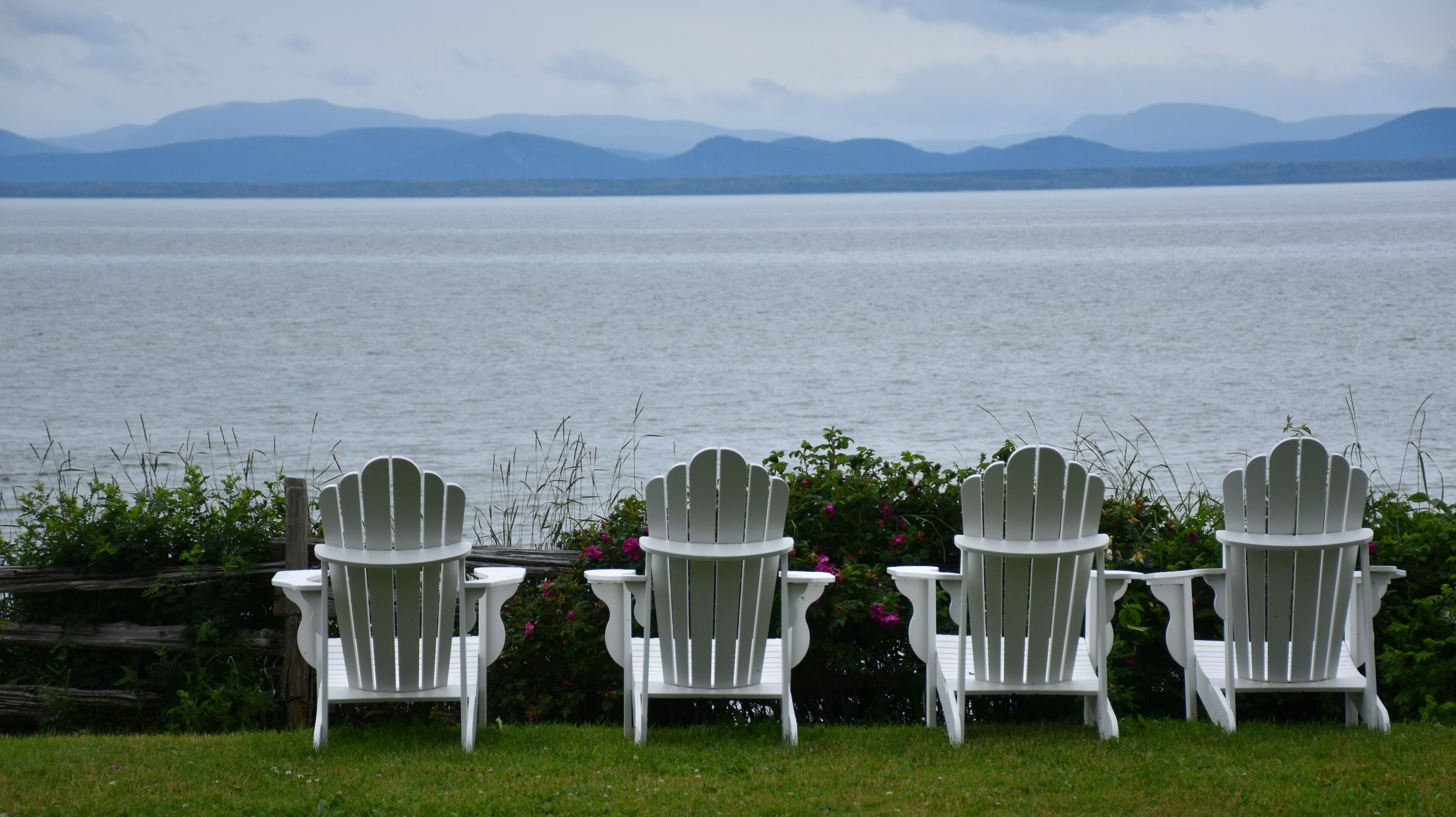 Three white Adirondack chairs facing a large body of water with mountains in the background.