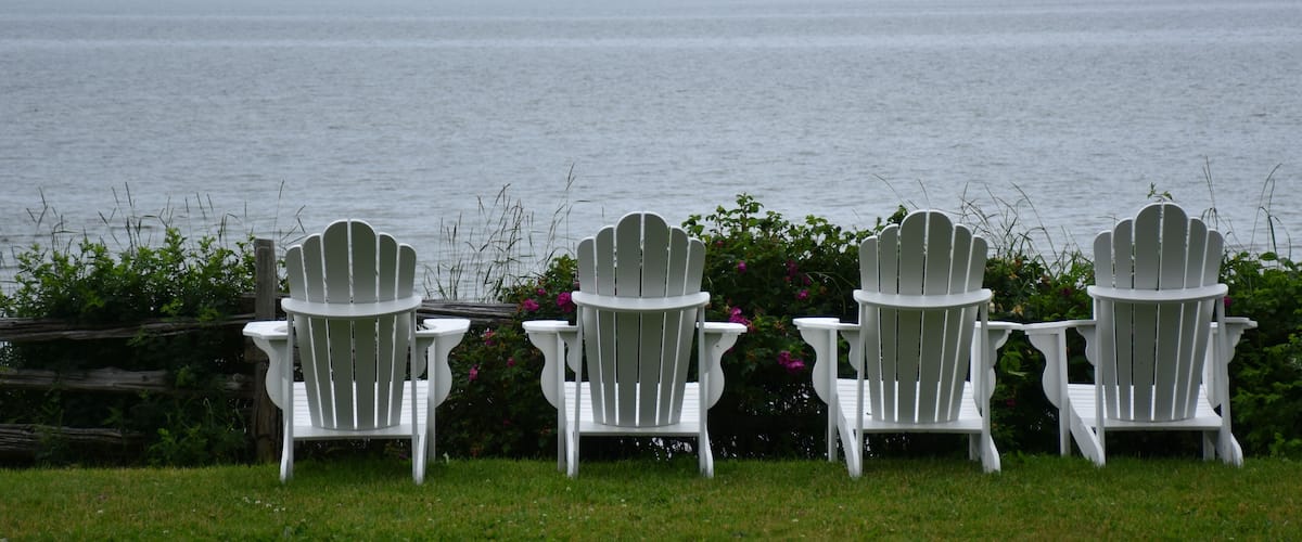 Three white Adirondack chairs facing a large body of water with mountains in the background.