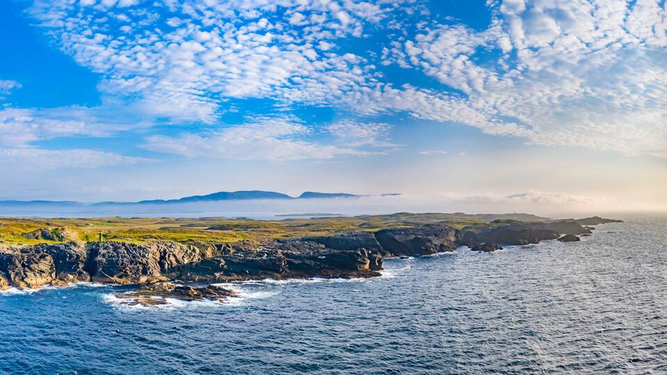 A coastal landscape with rocky cliffs, a calm sea, and a clear sky with scattered clouds.
