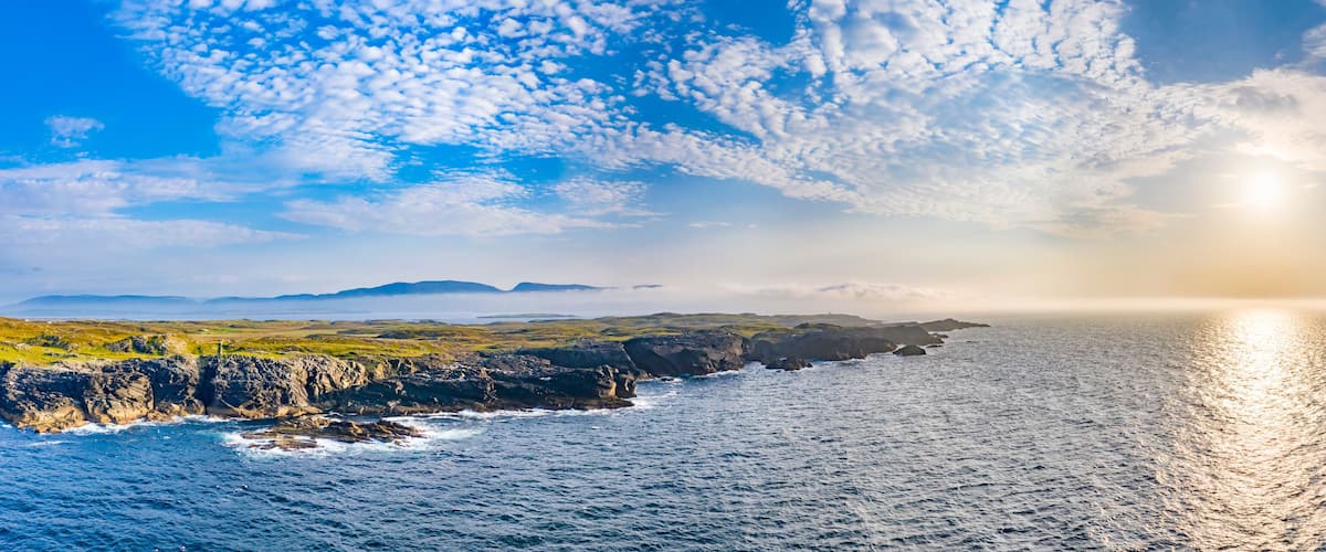 A coastal landscape with rocky cliffs, a calm sea, and a clear sky with scattered clouds.