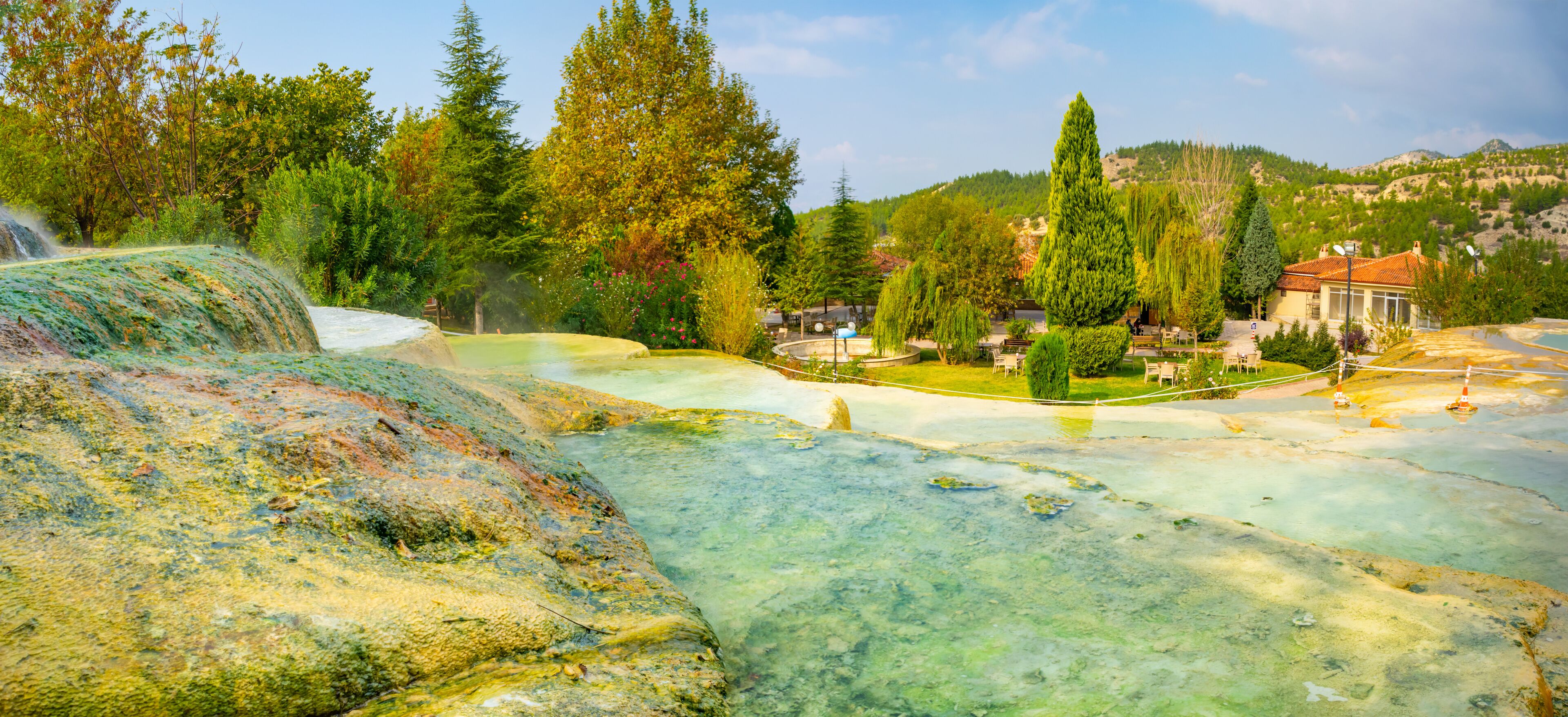 A geothermal area with mineral pools, trees, and a building in the background.