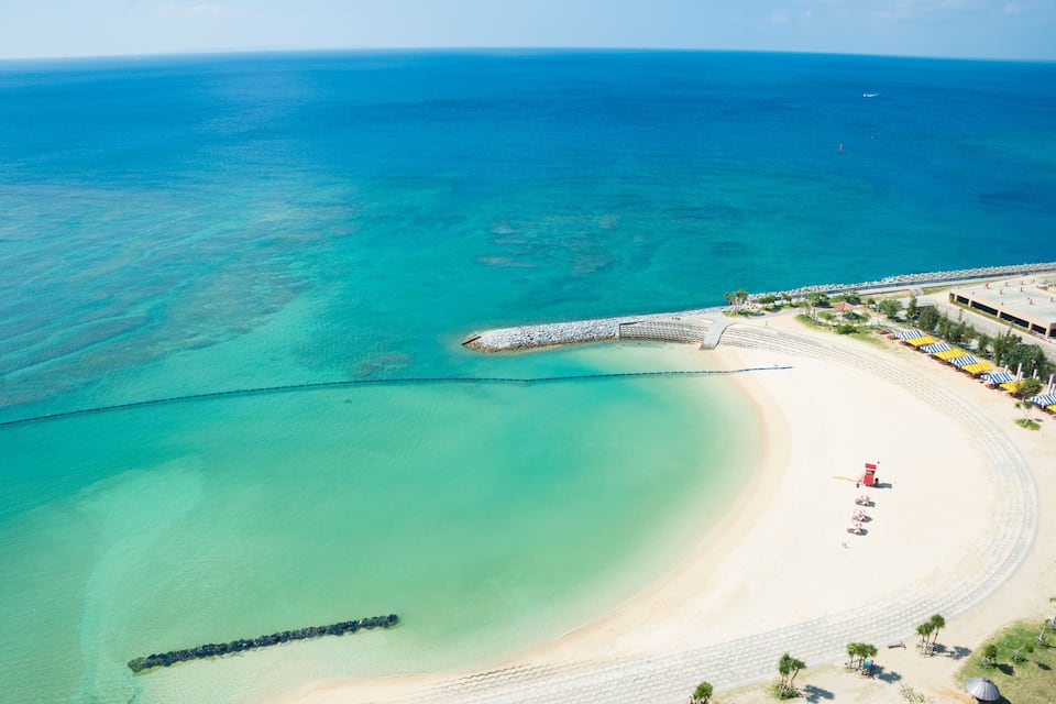 Une plage côtière aux eaux turquoise et limpides, avec un rivage sablonneux et une jetée qui s’avance dans la mer.