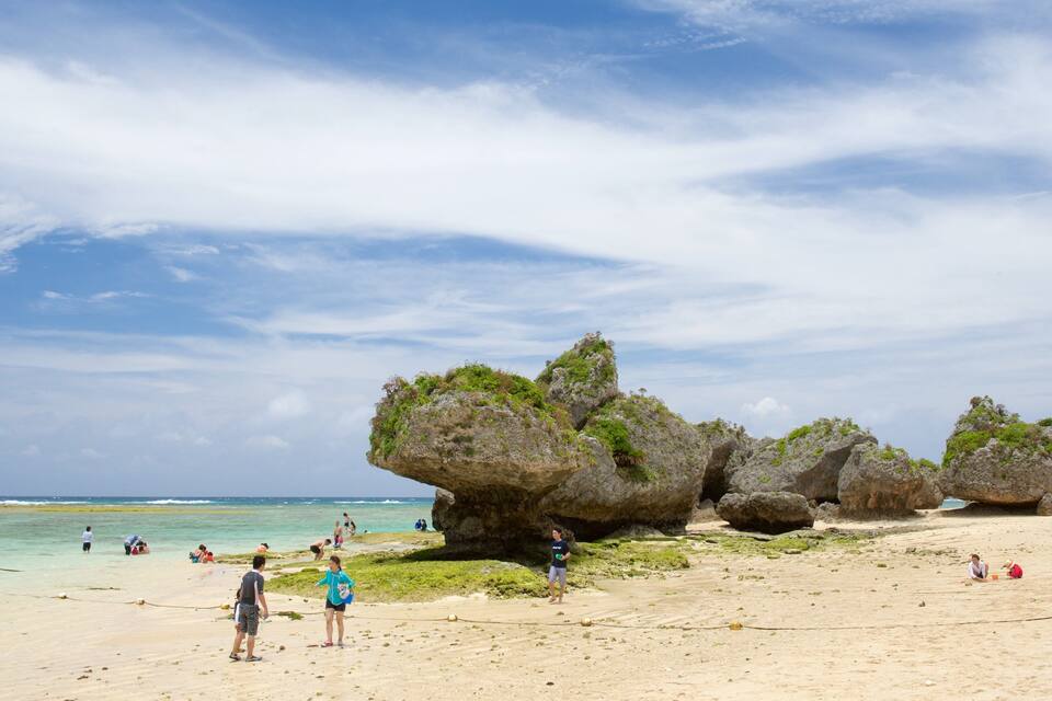 A beach with clear turquoise water, people sunbathing, and large rock formations.