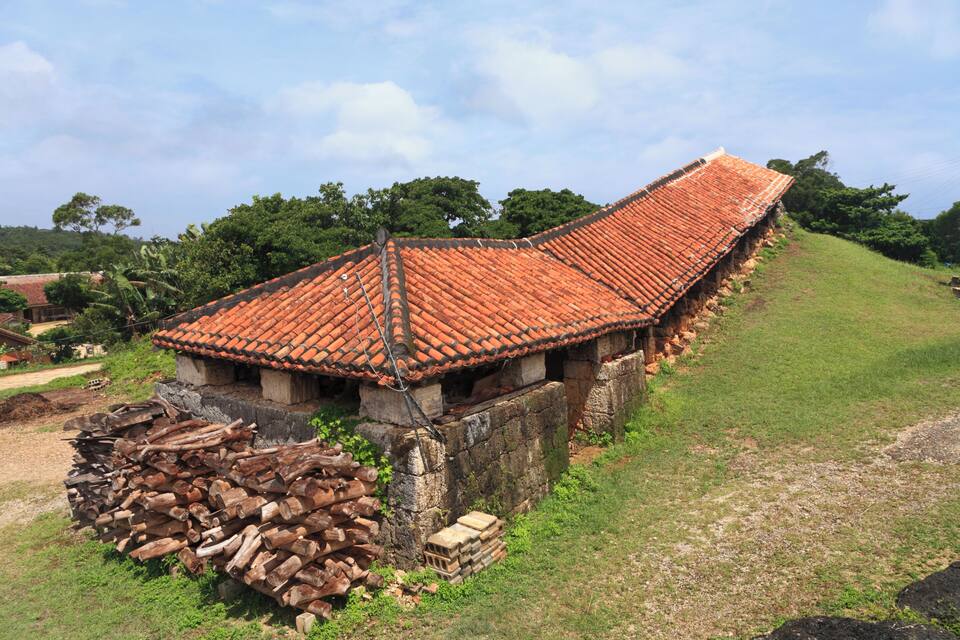 Une maison traditionnelle avec un toit à pente recouvert de tuiles rouges, entourée d’une colline herbeuse et d’arbres.
