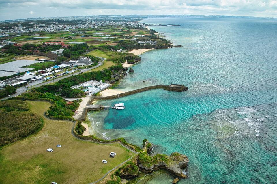 An aerial view of a coastal area with clear turquoise waters, a sandy beach, and a rocky outcrop.