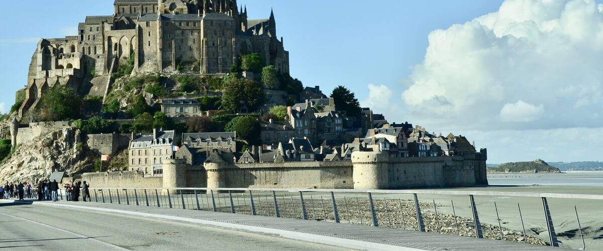 A historic castle on a hill with a prominent spire, surrounded by a fortified wall and a beach in the foreground.