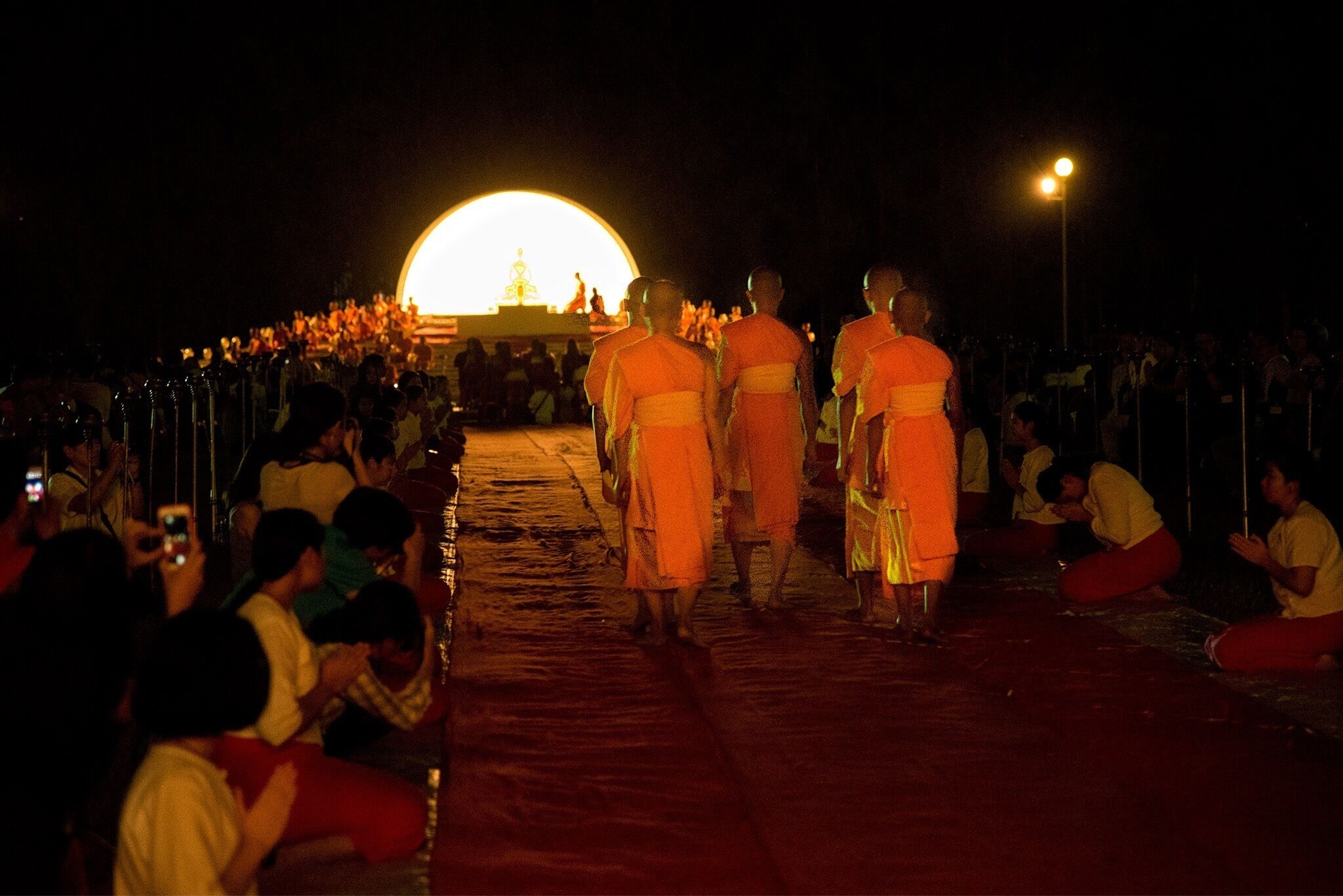 Monks walking down a street at night with a large illuminated structure in the background.