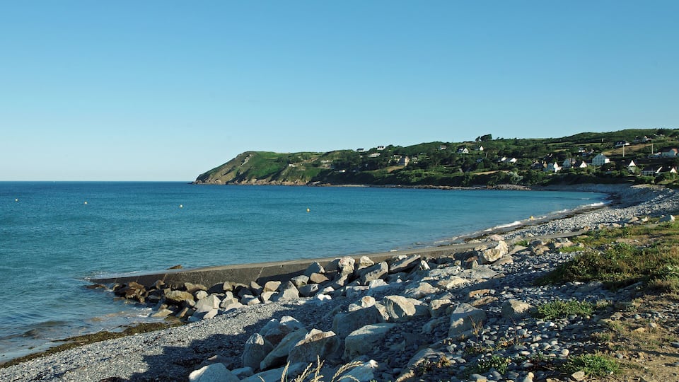 A rocky beach with a clear blue sea, a rocky breakwater, and houses on a hillside in the background.