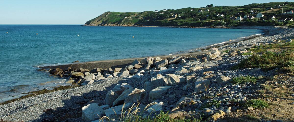 A rocky beach with a clear blue sea, a rocky breakwater, and houses on a hillside in the background.