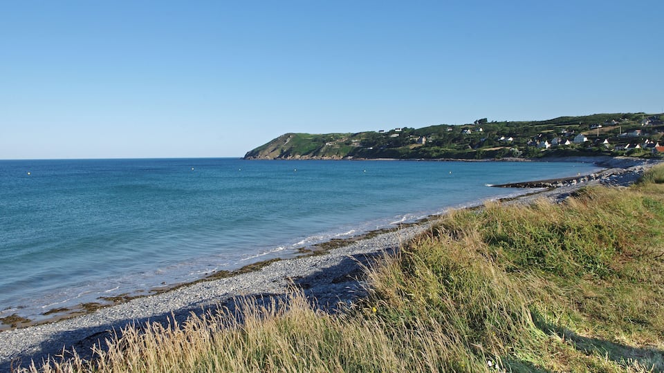 A coastal landscape with a pebbly beach, grassy dunes, and a hillside with scattered houses.