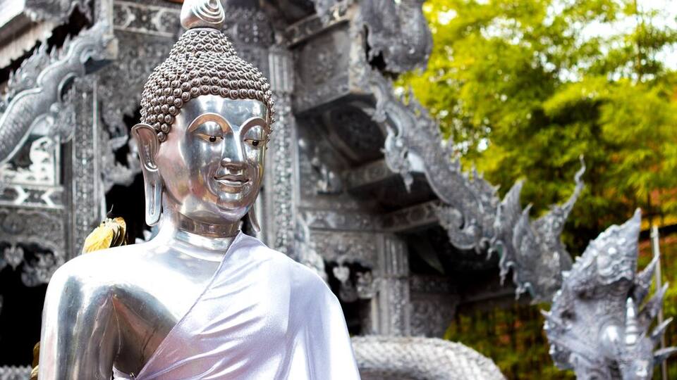A large, silver Buddha statue with intricate carvings, set against a backdrop of ornate temple architecture and lush greenery.
