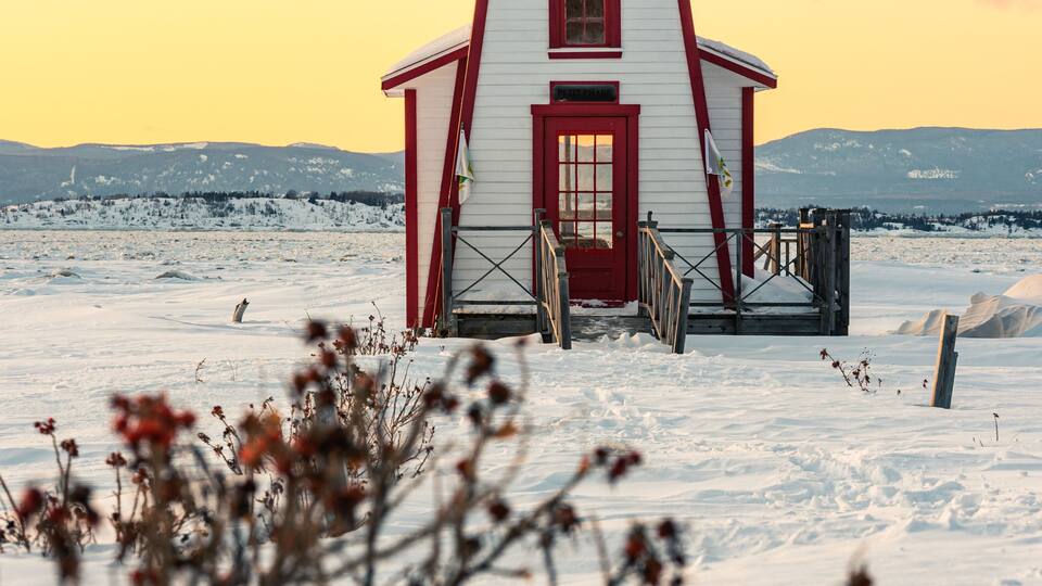 A red and white lighthouse in a snowy landscape with mountains in the background.