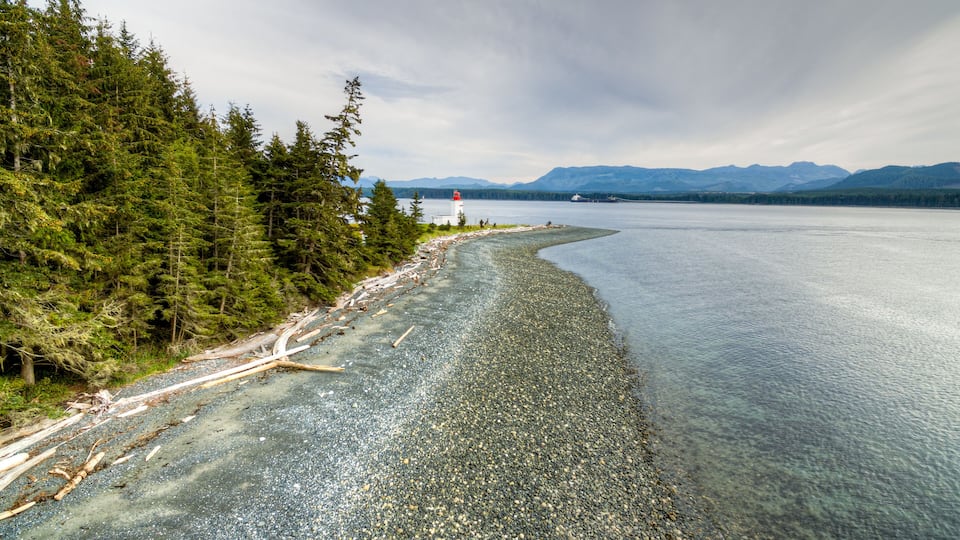 A pebble beach with a forest on one side and a lighthouse in the distance.
