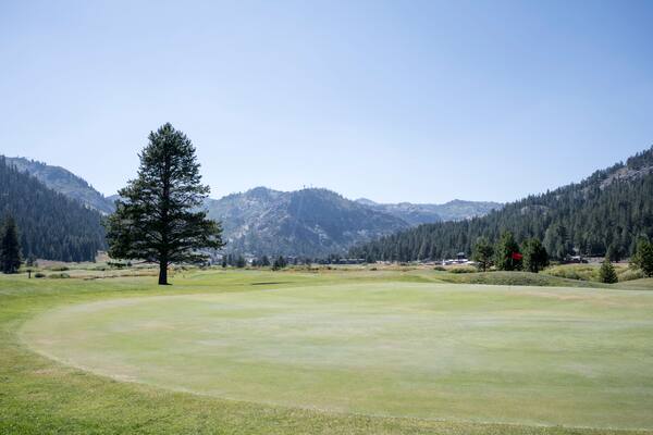 View across a large green on a mountain golf course. The mountains can be seen in the distance, against a blue, summers sky.