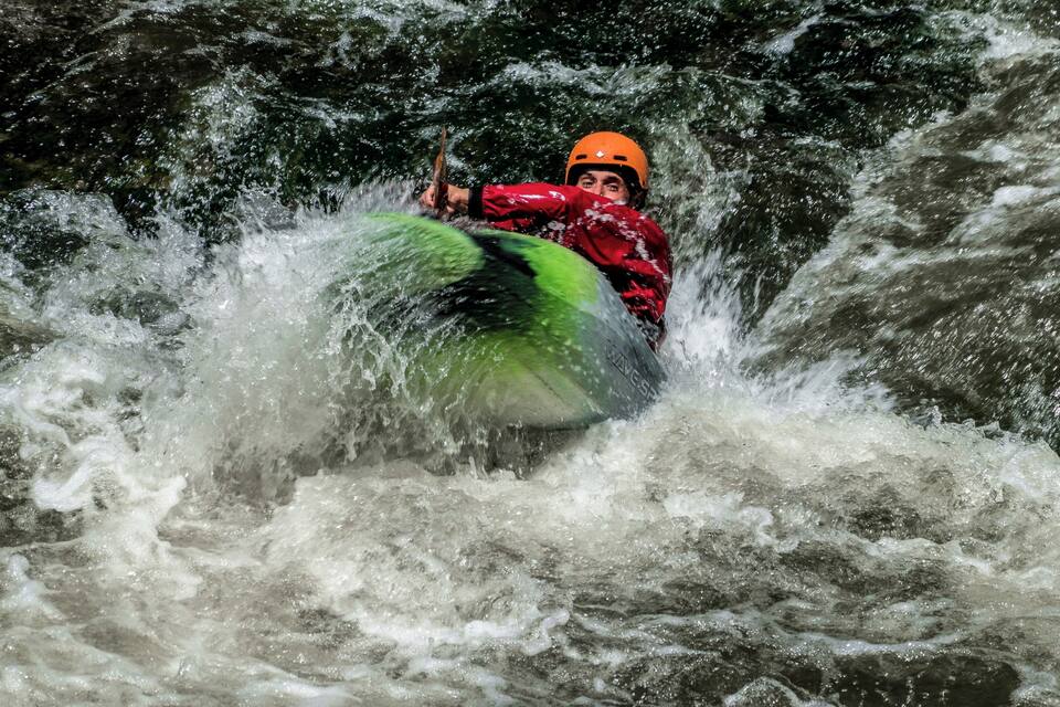 Une personne vêtue d’une veste rouge et d’un casque orange navigue sur un tronçon de rivière aux rapides tumultueux.