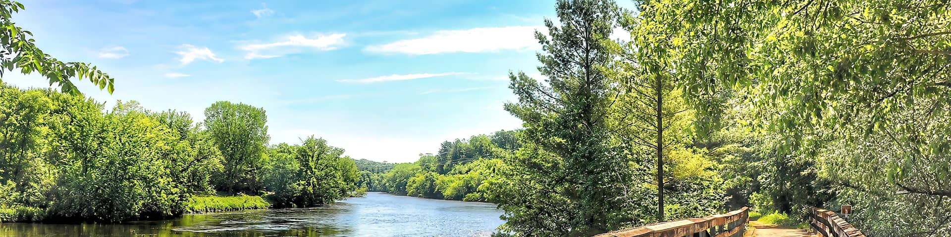 Beneath the blue sky and white clouds of a beautiful summer day in Wisconsin, the Red Cedar State Bike Trail crosses a wooden bridge and passes along the Red Cedar River through a lush green forest.