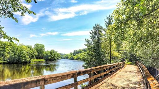 Beneath the blue sky and white clouds of a beautiful summer day in Wisconsin, the Red Cedar State Bike Trail crosses a wooden bridge and passes along the Red Cedar River through a lush green forest.