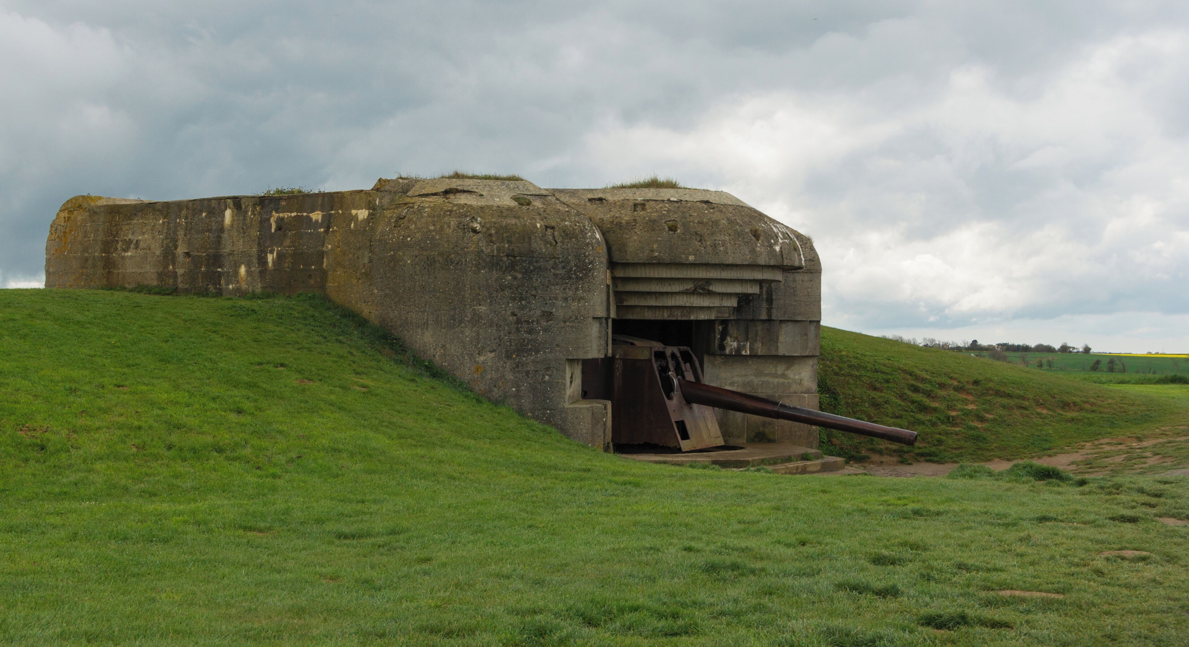 A large concrete bunker with a gun barrel protruding from the front, situated on a grassy hill under a cloudy sky.