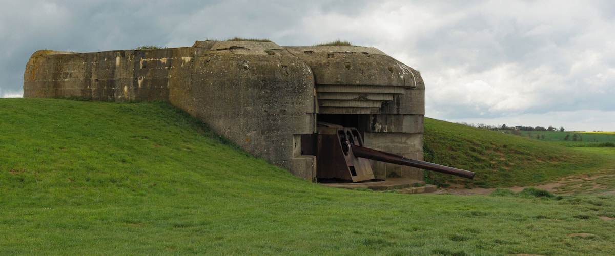 A large concrete bunker with a gun barrel protruding from the front, situated on a grassy hill under a cloudy sky.