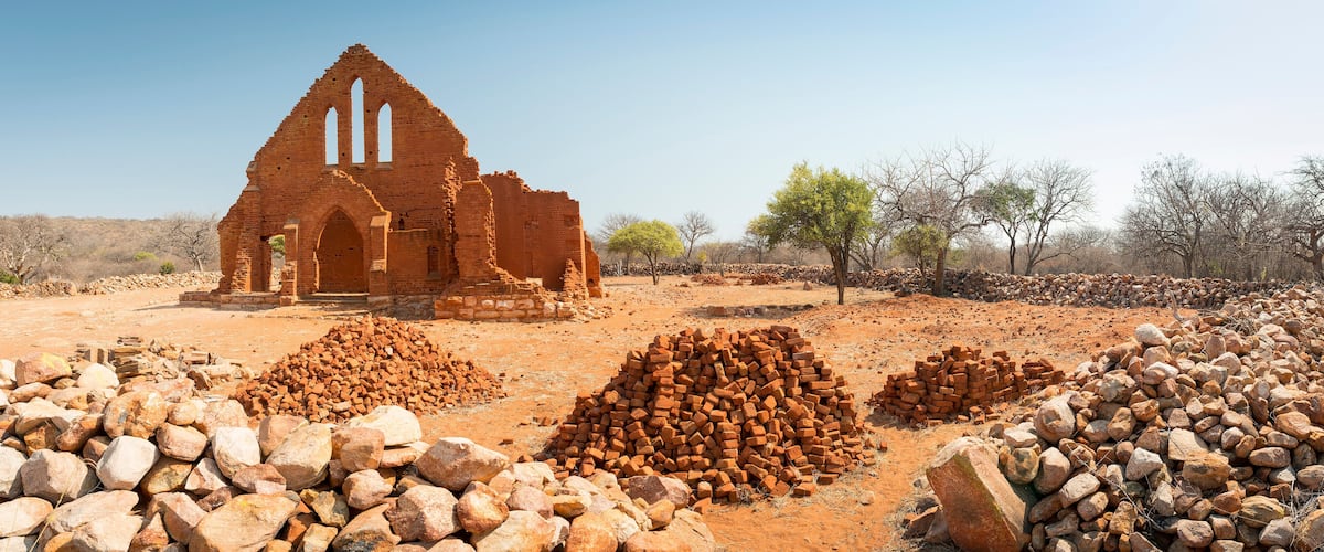 An ancient brick church with a triangular roof and arched doorway, surrounded by piles of bricks and stones in a dry, open landscape.
