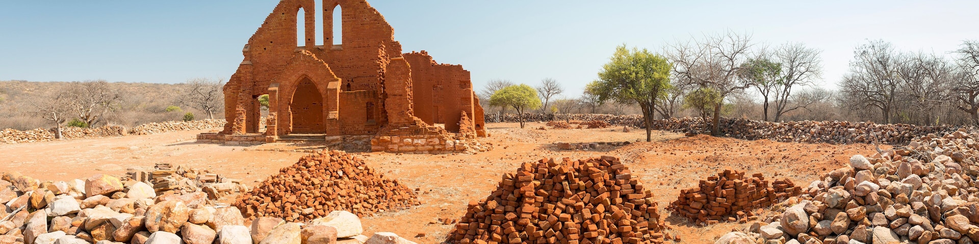 An ancient brick church with a triangular roof and arched doorway, surrounded by piles of bricks and stones in a dry, open landscape.