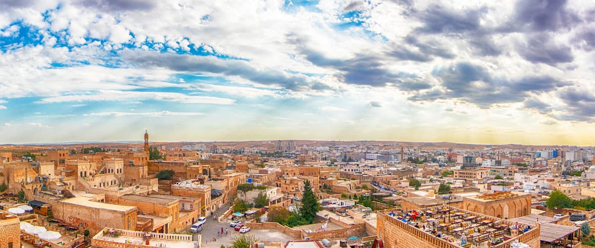 Panoramic view of Midyat in Mardin, Turkey, showcasing historic stone buildings, traditional architecture, and a vast cityscape under a dramatic cloudy sky.