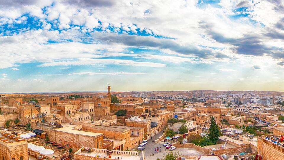 Panoramic view of Midyat in Mardin, Turkey, showcasing historic stone buildings, traditional architecture, and a vast cityscape under a dramatic cloudy sky.