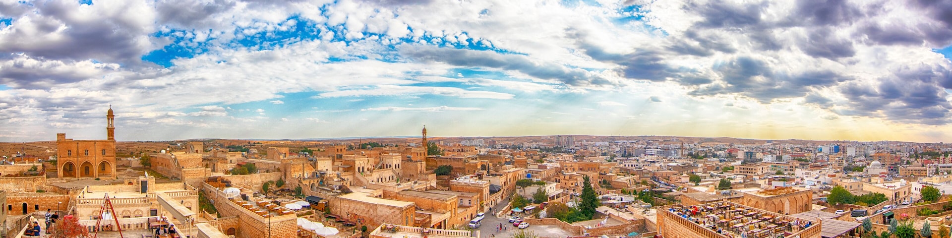 Panoramic view of Midyat in Mardin, Turkey, showcasing historic stone buildings, traditional architecture, and a vast cityscape under a dramatic cloudy sky.