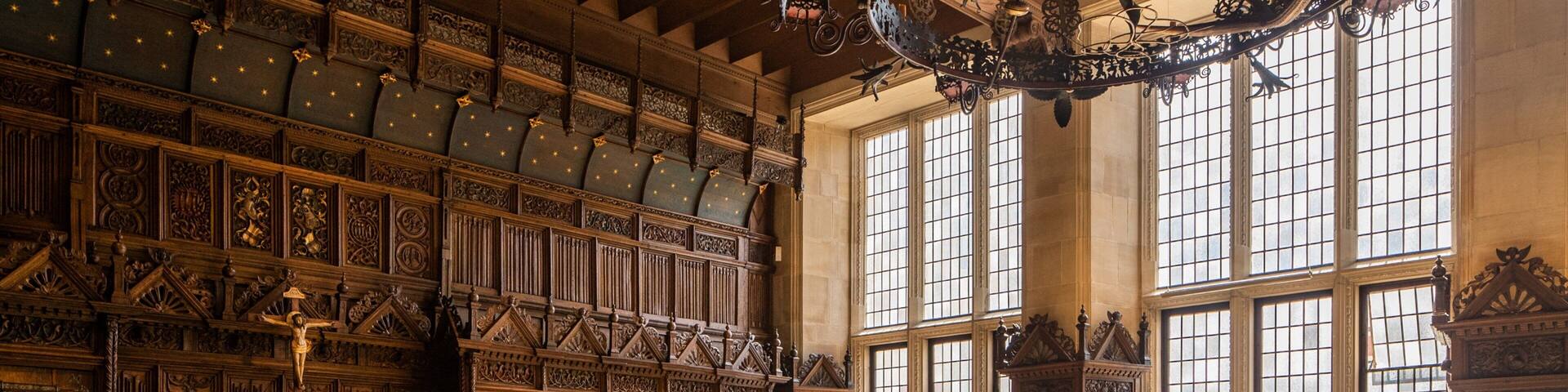 An ornate wooden room with intricate carvings, large windows, and a chandelier.
