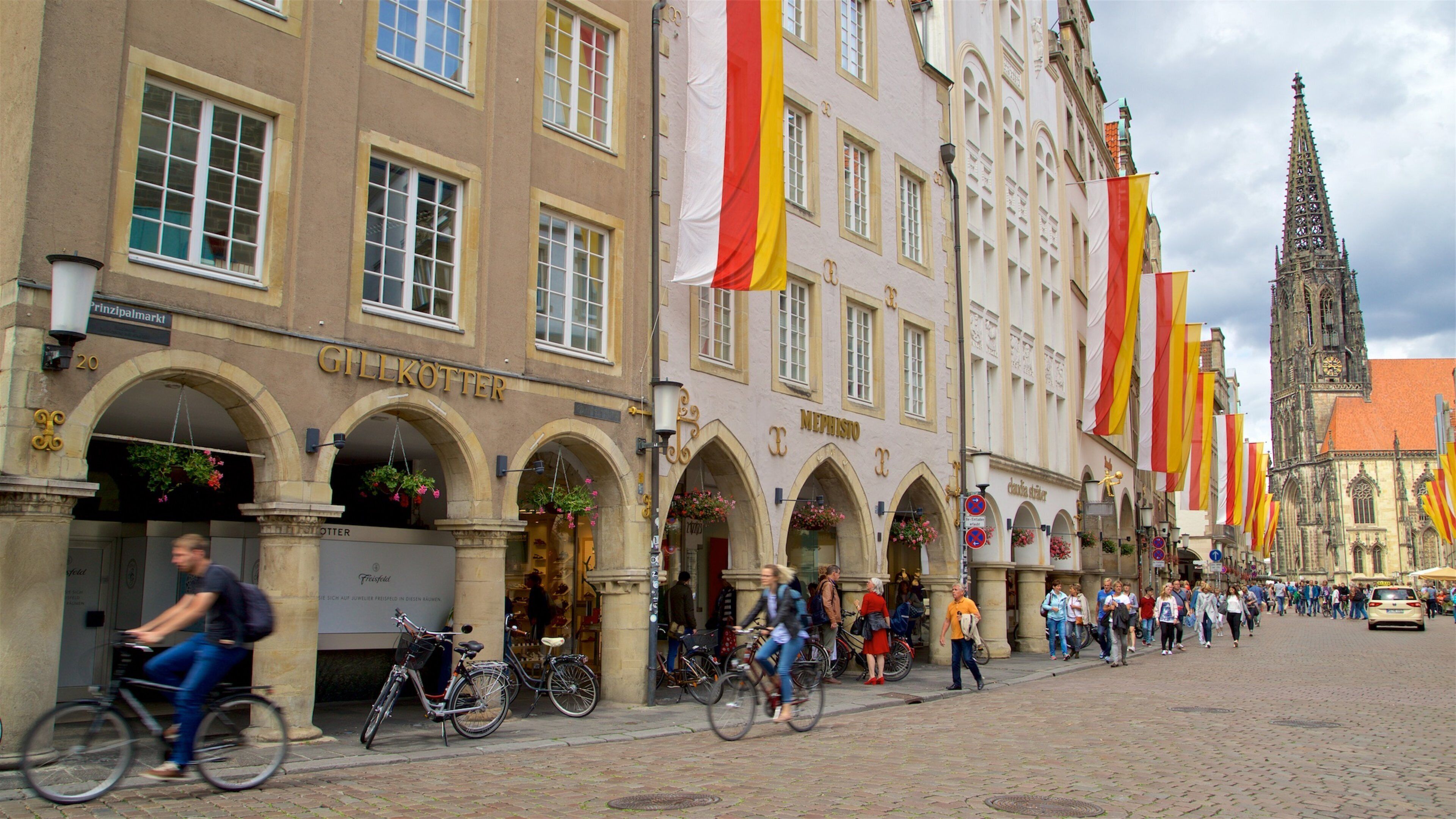 Eine Kopfsteinpflasterstraße gesäumt von historischen Gebäuden, darunter ein Geschäft namens "GILLKOTTER" und eine Kirche mit einem hohen Turm.