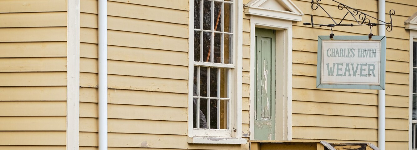 A yellow building with a sign reading 'Charles Irvin Weaver,' a wooden staircase, and a weathered fence.