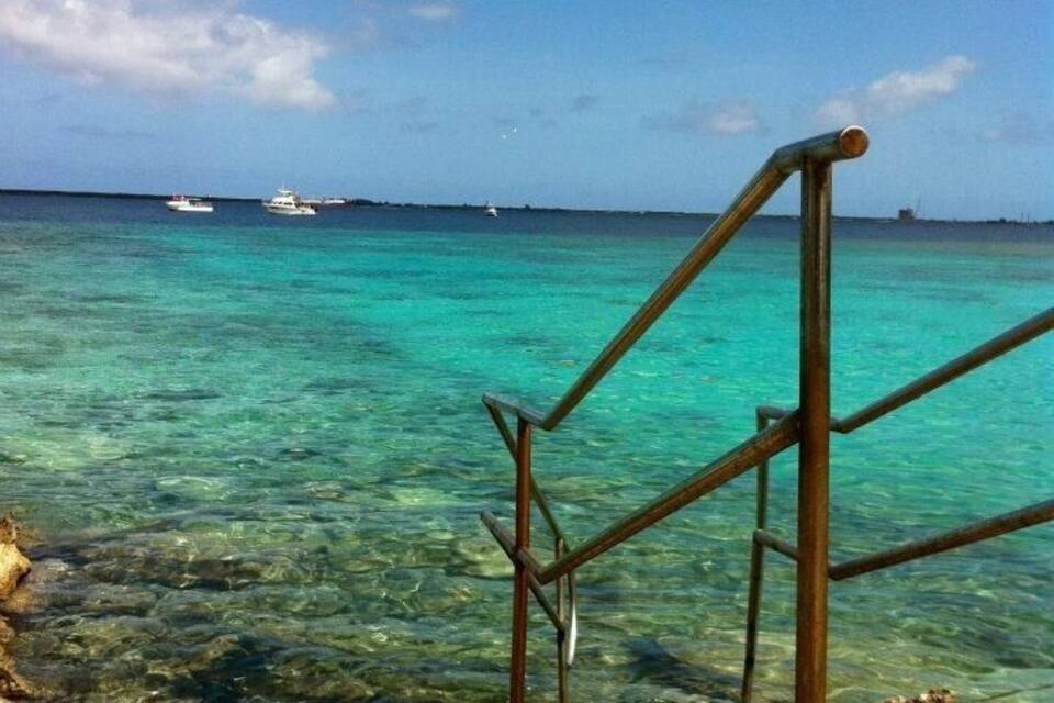 A clear turquoise sea with a metal staircase leading down to the water, boats in the distance, and a clear sky.