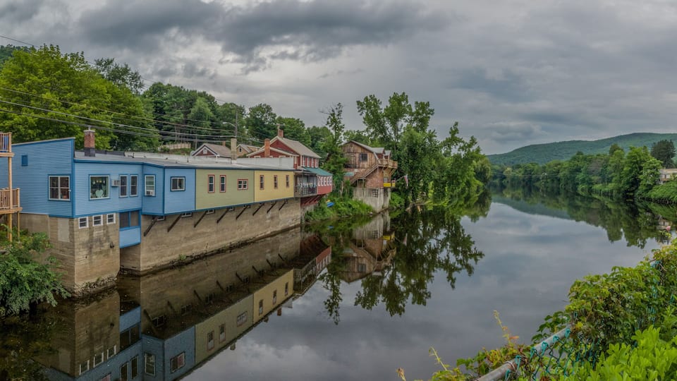 A river with buildings reflected on the water, surrounded by greenery and hills.
